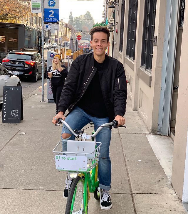 William Givens Jensen in a black t-shirt, blue jeans, and black jacket riding a bicycle. 
